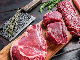 Fresh cuts of raw beef displayed on a wooden board with herbs nearby, illustrating the ongoing debate about whether dogs can eat raw beef or if raw food diets are beneficial, as many pet owners consider raw feeding for their dogs despite veterinary discussions about the safety of uncooked meat.
